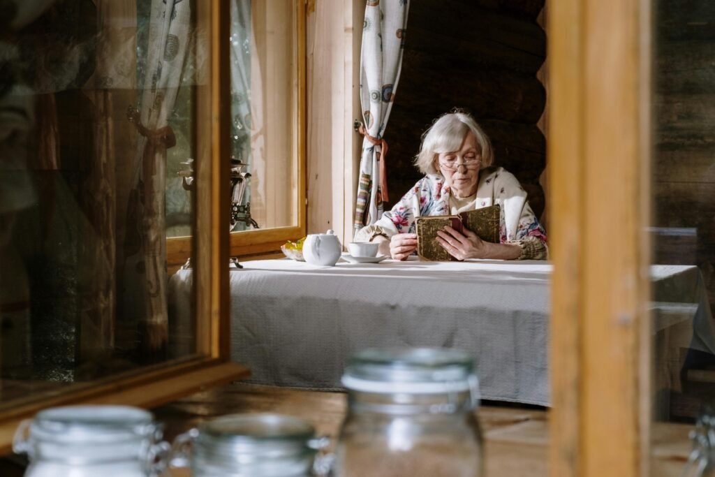 Senior woman reads by the window in a cozy cabin, enjoying a cup of tea with a teapot on the table. Ins and Outs 2025.