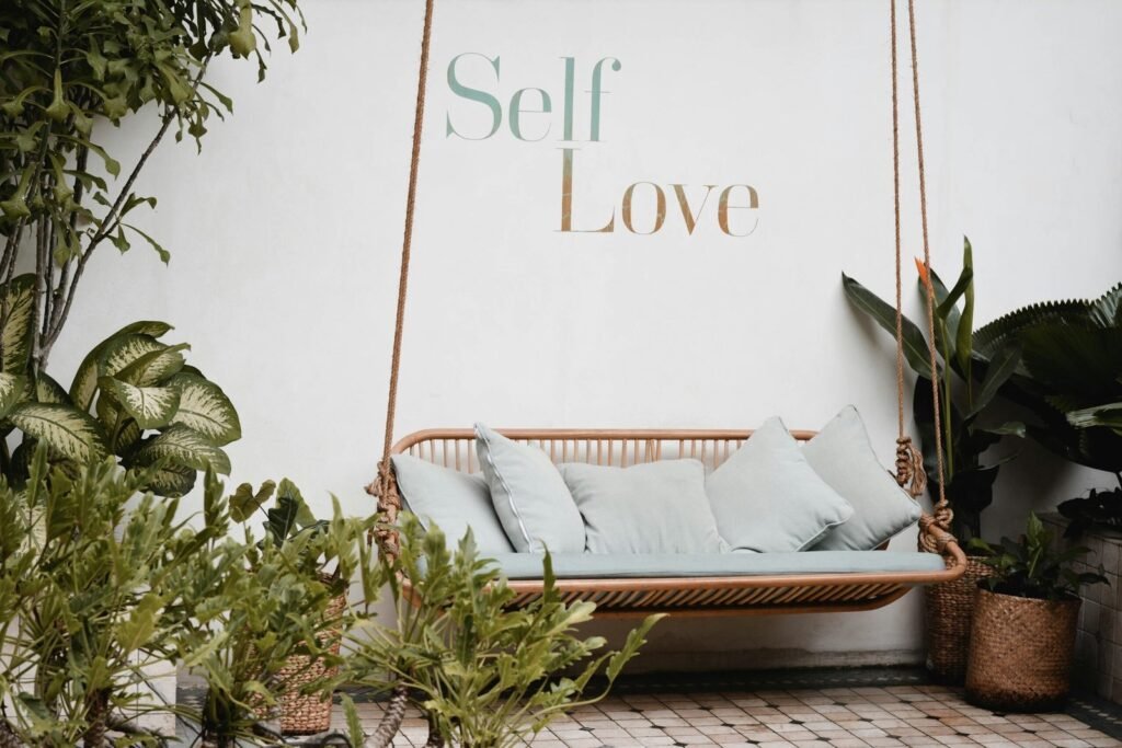 the words self love written on a white wall with a hanging love seat in front of it, surrounded by indoor greenery